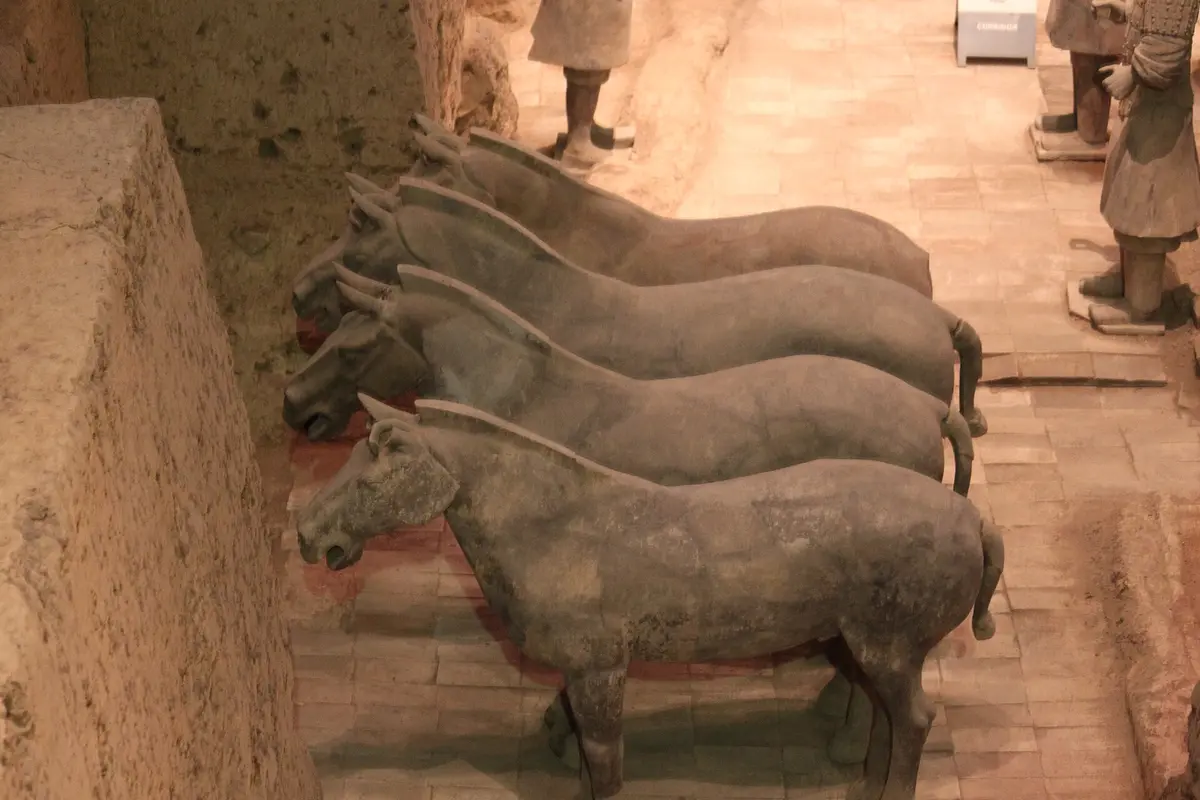 Four terracotta horses standing side by side in a pit, with a terracotta figure at the edge of the frame